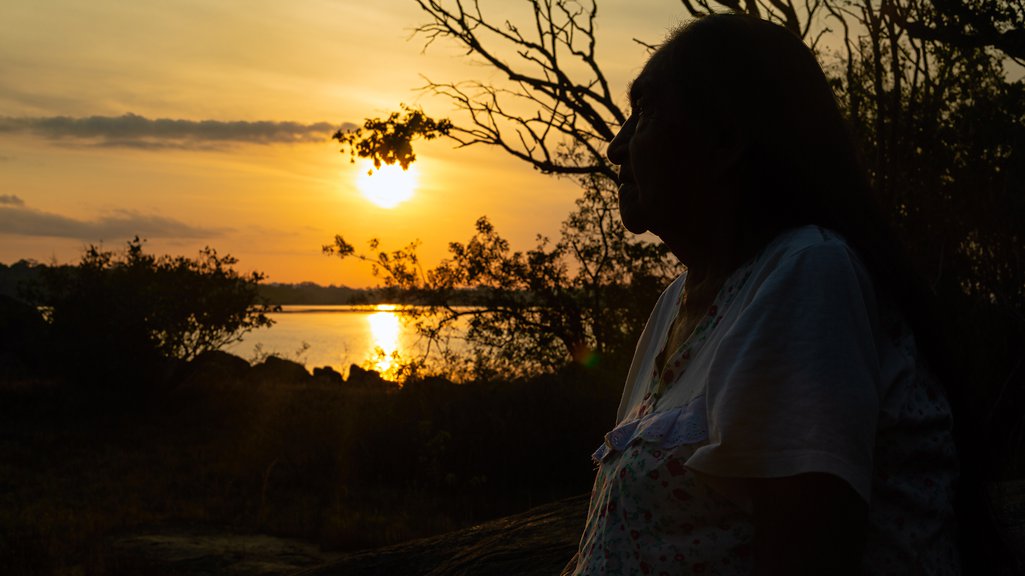 Odete Kuruayade perfil em frente a um lago durante o pôr-do-sol