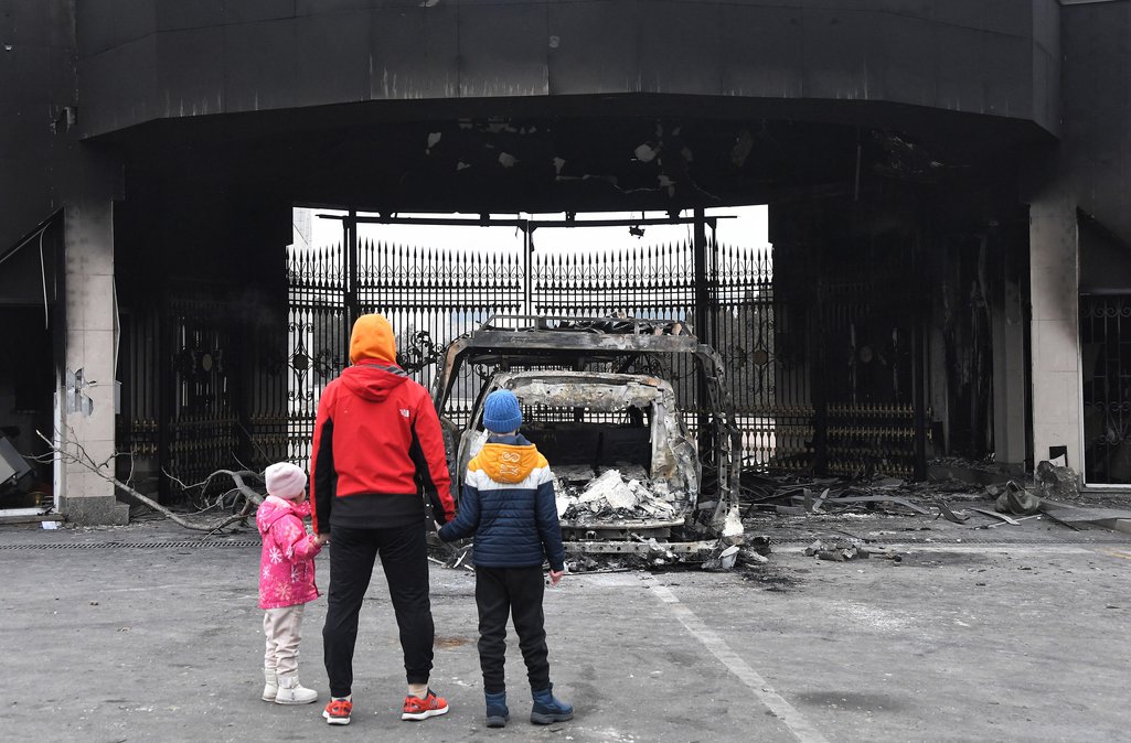 Two children hold hands with an adult in front of a burned-out car on a Kazakhstan street