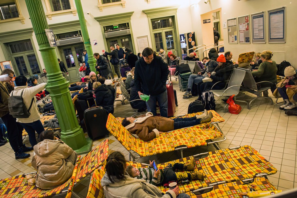 Pessoas deitadas em camas de acampamento em uma estação de trem lotada, cercadas por bagagens