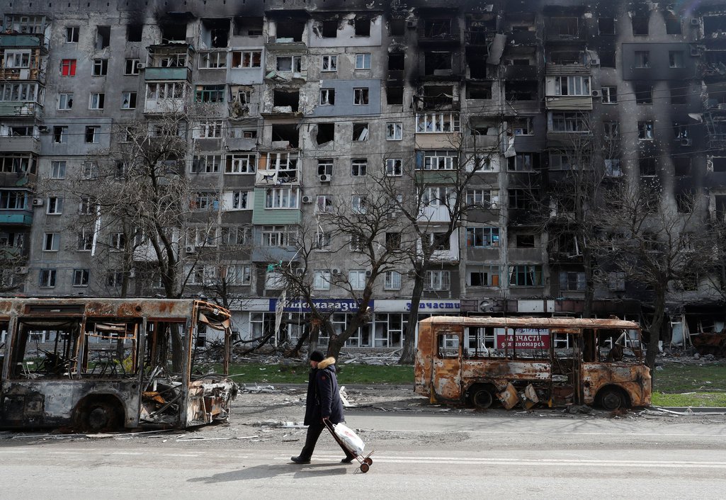 A Mariupol resident walks past burnt-out buses in front of a heavily shelled apartment block