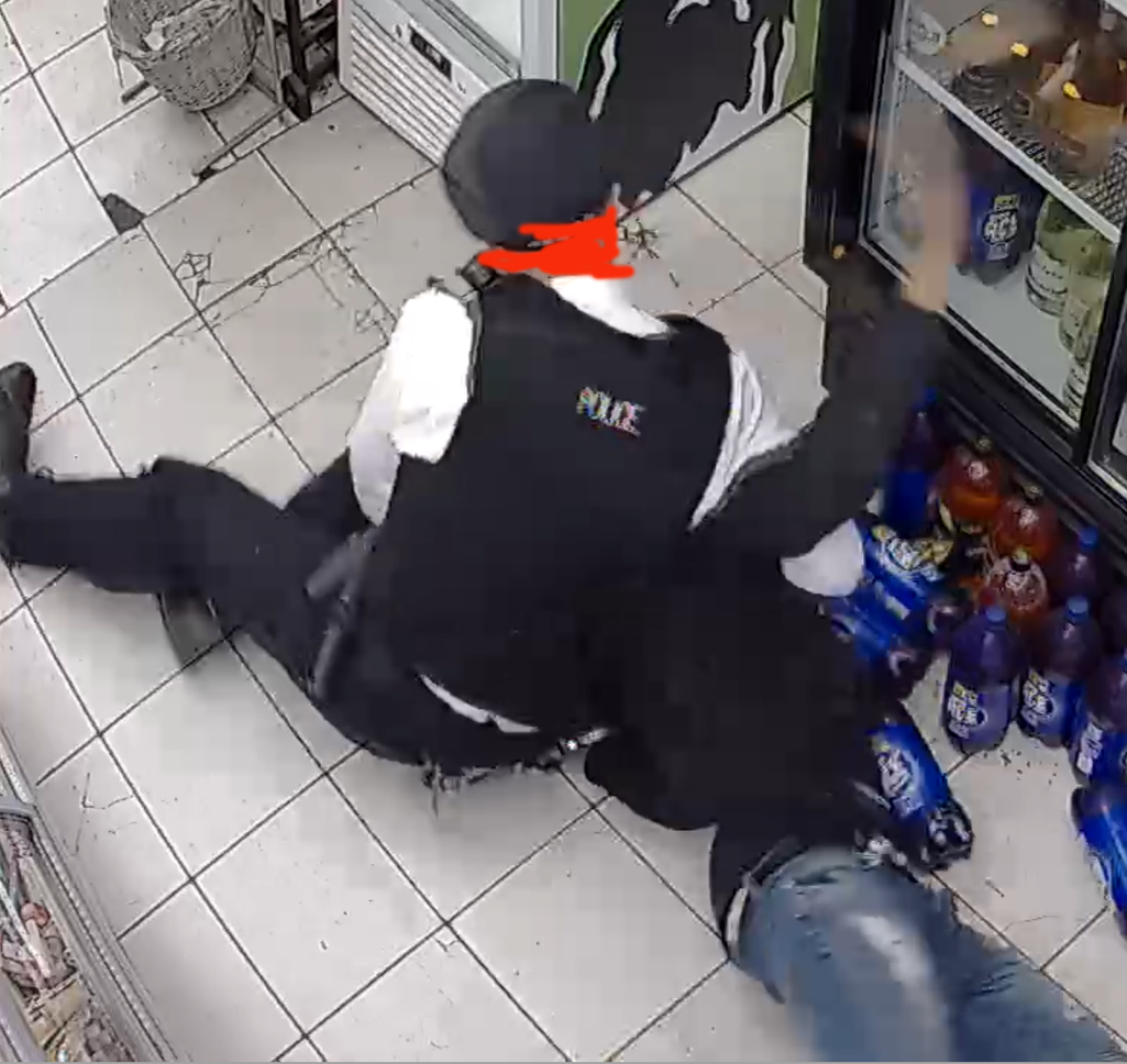Police officer pins down young man, whose hand is tapping against tall commercial drinks fridge