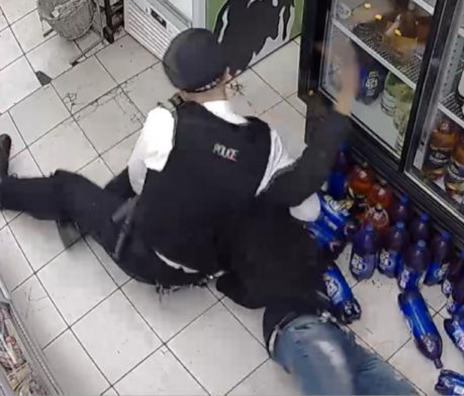 Police officer holds young man, their faces are obscured. The young man's hand taps a drinks fridge. They are inside a small shop.