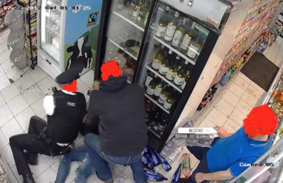 One man observes two men restraining a young man on the floor of a small shop.