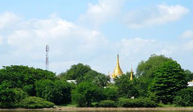 A mobile phone antenna and golden temple behind green trees alongside a river