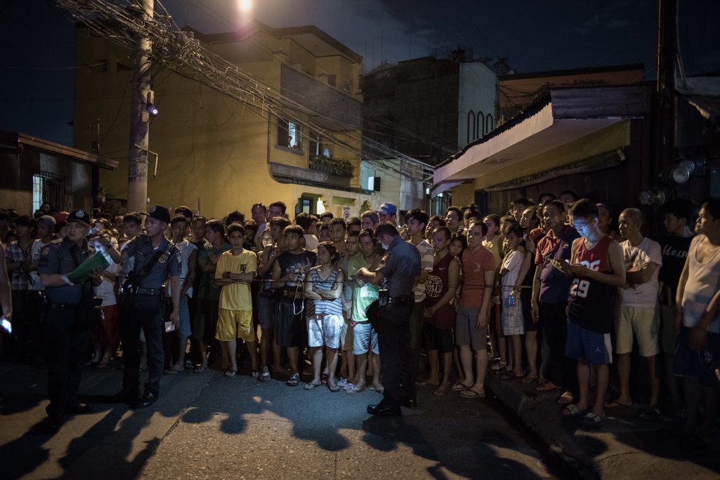 A crowd of people behind a police cordon at night in a city street.