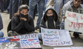 Two women sit with signs and symbolically gagged mouths alongside a crouching man with a sign.