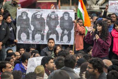 A young women speaks into a mic to a large crowd. Poster of the three wise monkeys behind, plus a 4th covering genitals.