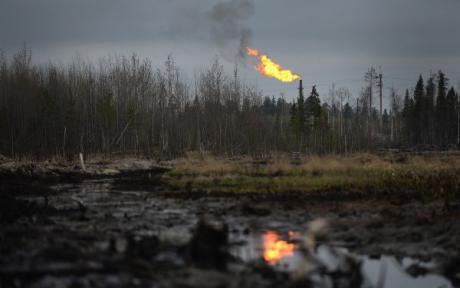Crude oil seeps through the ground, in the background, a derrick burns off gas.