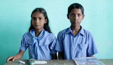 Bangladeshi schoolgirl and schoolboy sit side-by-side at a school desk: looking straight at the camera.