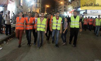 A group of 7 or so men in high-visibility vests walk down the middle of a street, stared at by on-lookers.