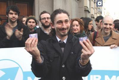 Man holds up two halves of a destroyed credit card, surrounded by campaigners
