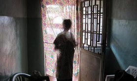 Woman stands at a window, silhouetted against curtain.