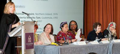 Six women seated on a panel, one standing at a podium.
