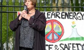Women stands speaking, microphone in hands, next to a peace sign .