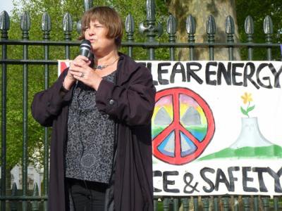 Women stands speaking, microphone in hands, next to a peace sign .