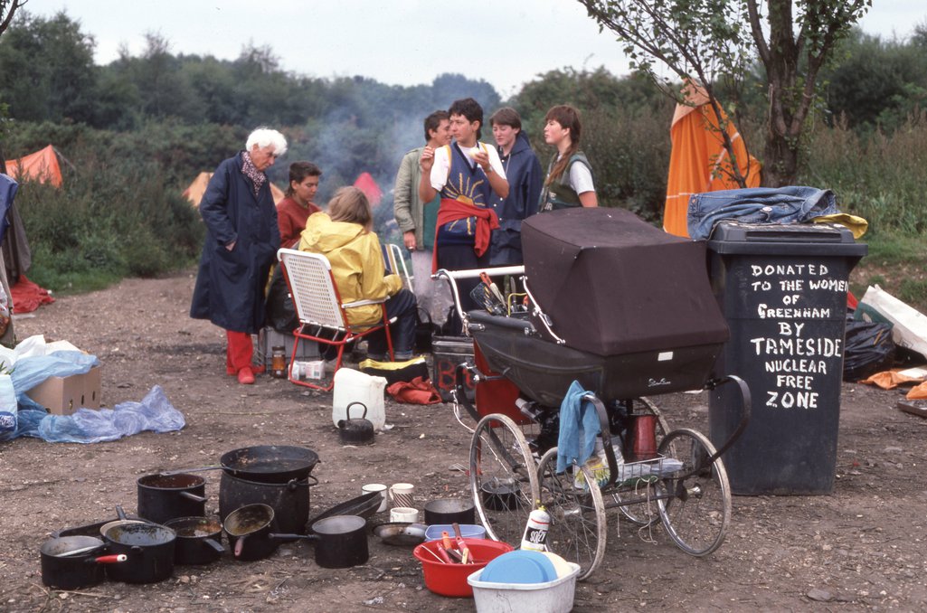 Women sit around a fire, in a clearing. Cooking utensils surround them and there is a pram filled with food