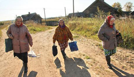 Three old Russian women walk down a dirt road in rural Russia carrying heavy bags. 