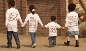Four children holding hands walking away from the camera, wearing T-shirts with Arabic text on.