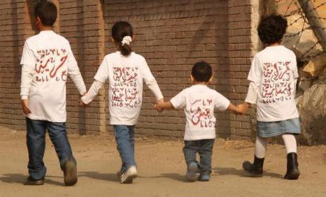 Four children holding hands walking away from the camera, wearing T-shirts with Arabic text on.