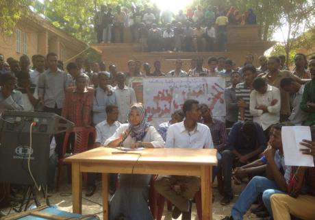Male and Female student sit at a desk outside in front of a camera. Hundreds of student supporters  look on.