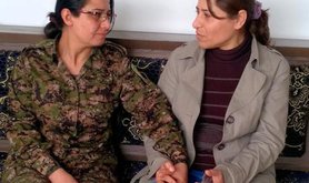 Two women sit holding hands, one of them in combat fatigues.