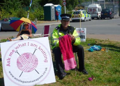 Policeman sits knitting a pink wool piece next to a Wool Against Weapons sign.