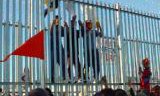 Three people climbing a high, razor-wire-topped fence. Red flag in the foreground.