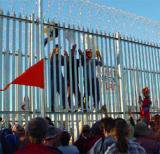 Three people climbing a high, razor-wire-topped fence. Red flag in the foreground.