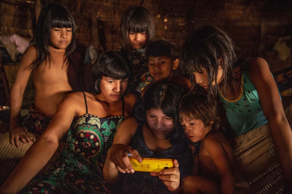 Um grupo de meninas e jovens indígenas contempla imagens em um telefone celular em sua aldeia