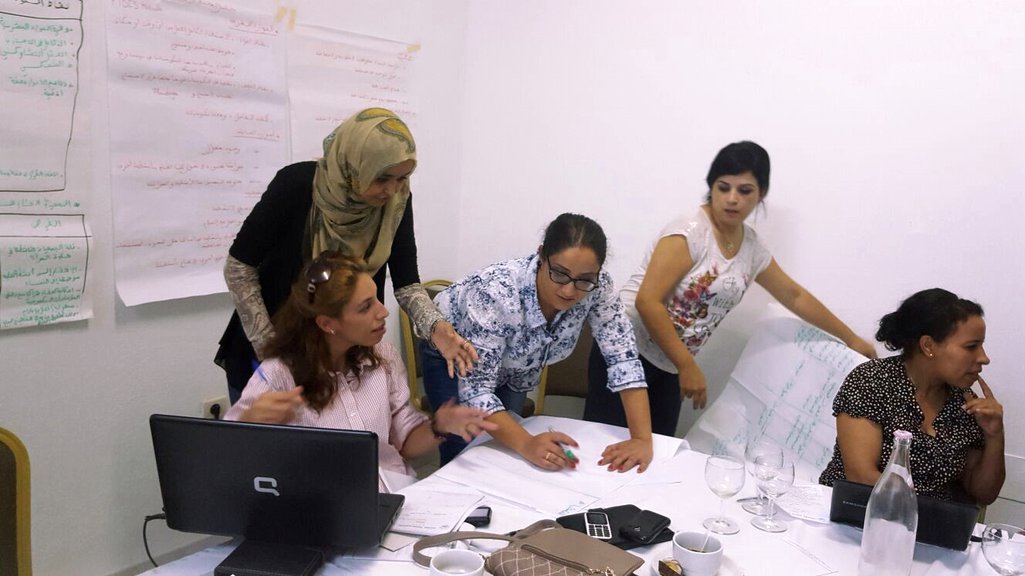 Women discussing things in a classroom with sheets of paper covered in Arabic writing on the walls