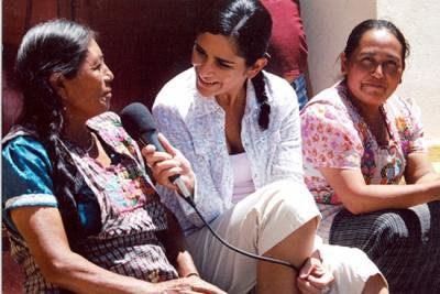 Woman with microphone sits on steps between two women in bright sunlight