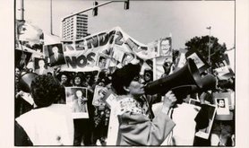 Black and white photo of street protest. Woman with megaphone and photograph, banners and placards in background..