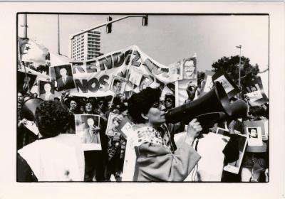 Black and white photo of street protest. Woman with megaphone and photograph, banners and placards in background..