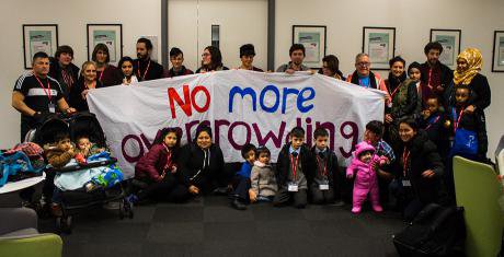 People of all ages including babies huddled together. They hold a large banner: no more overcrowding.