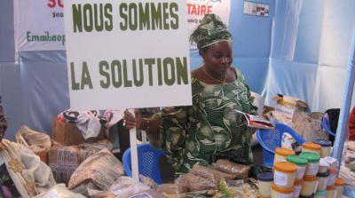 Woman at a market stall holding a placard saying 'Nous sommes la solution'