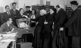 Black and white archive image of 1930s polling station with 4 women lined up to vote.